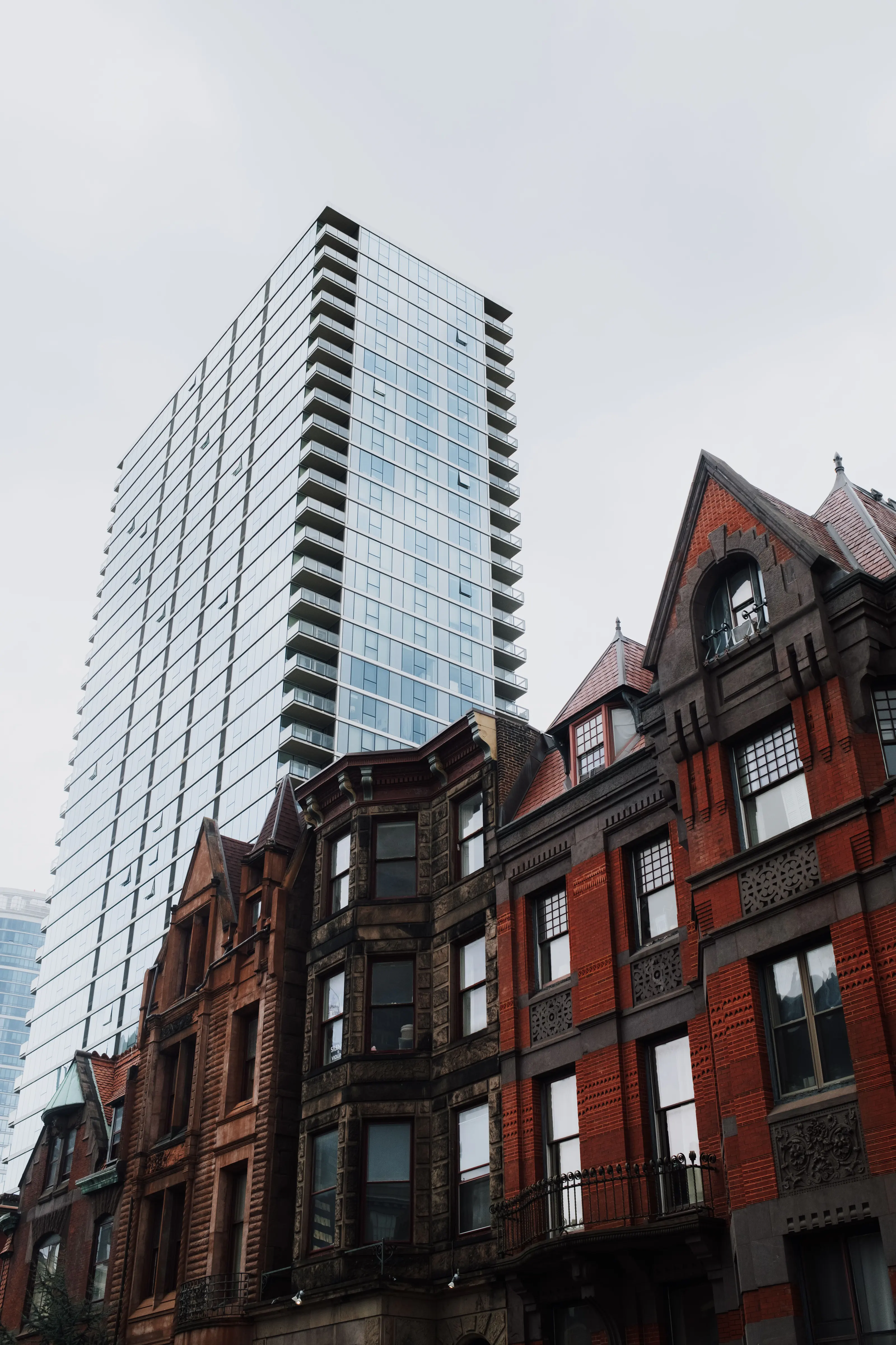 A glass residential tower rising directly behind a terrace of Victorian brick and greystone houses with steep gables and bay windows, under a flat pale sky