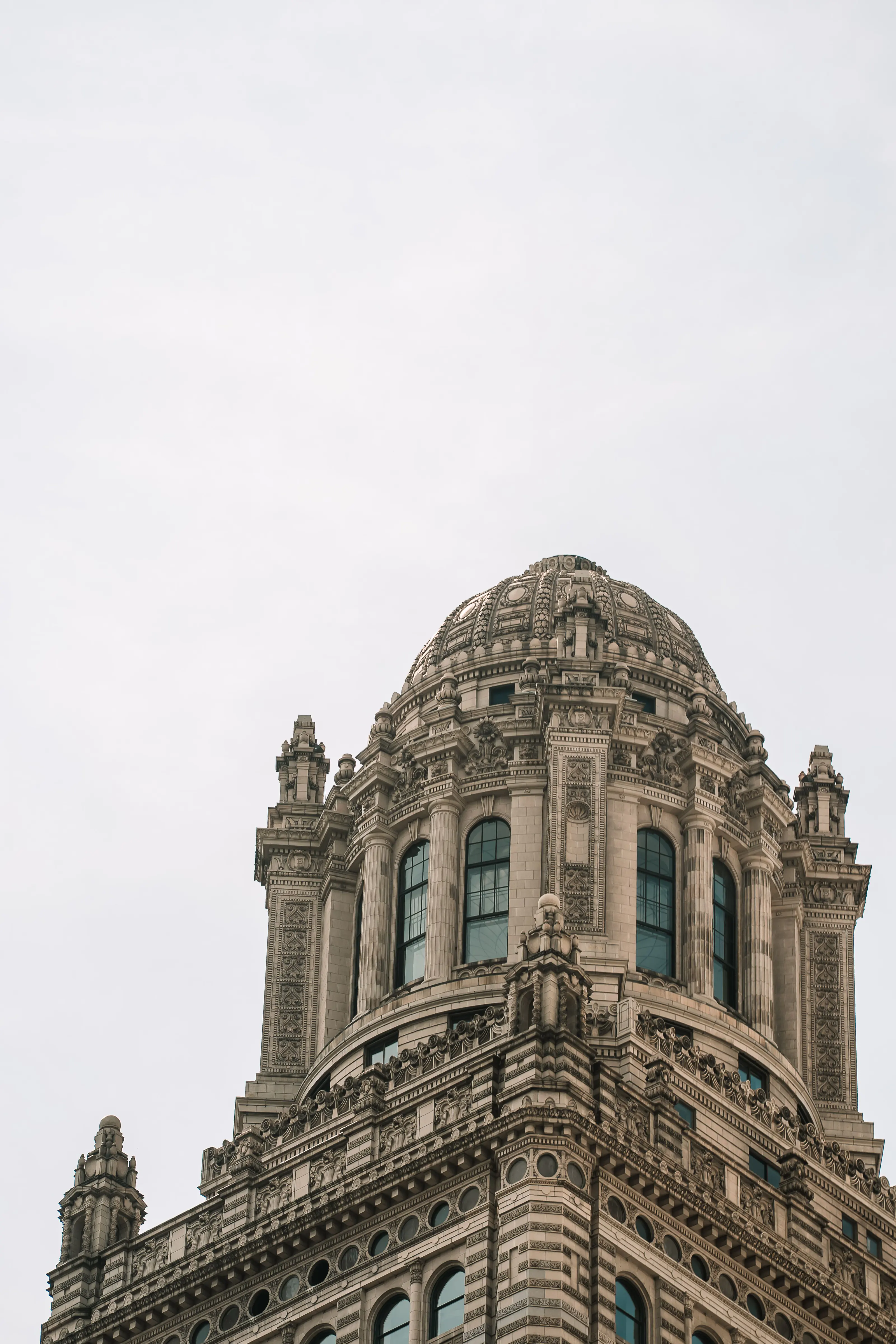 The crowning storeys of a classical Chicago tower — a carved masonry dome above an arcade of columns, flanked by corner turrets — against a flat pale sky
